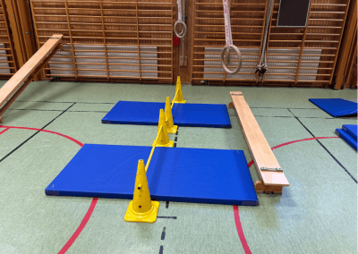 Hinderbana i gympasal – En station med ringar och hinder som tränar styrka, koordination och kroppskontroll. Barnen kan svinga sig i ringarna och landa på mattan, eller försöka ta sig över ett hinder.
