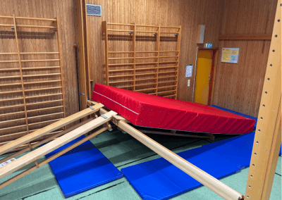 Hinderbana i gympasal – En station med lutande plint och madrass som tränar klättring, koordination och mod. Barnen kan klättra uppför rampen och ta sig över madrassen.